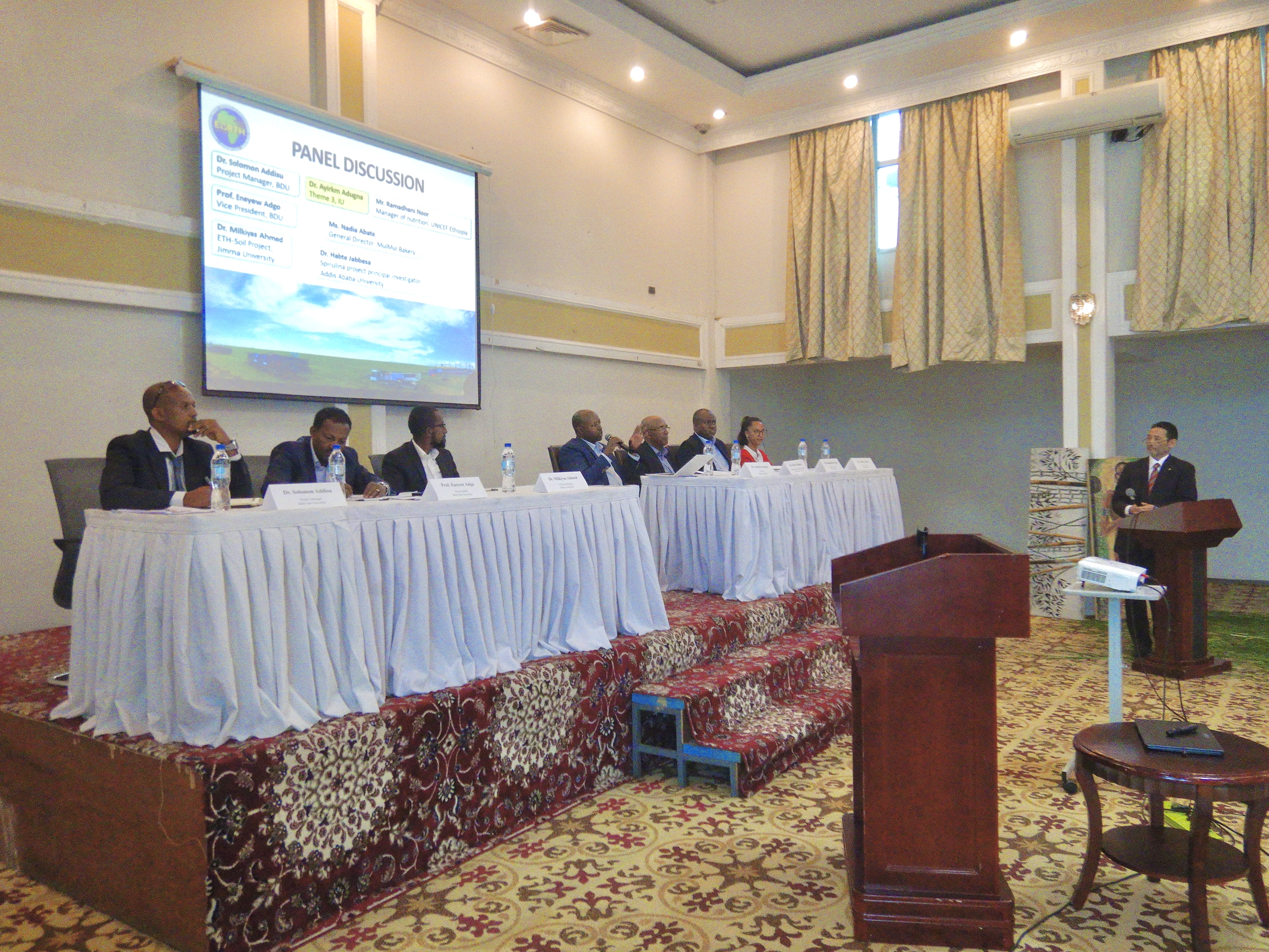 ステージに7名のパネリストが横並びに座っている。ステージ下の演台から佐藤教授が質問している。