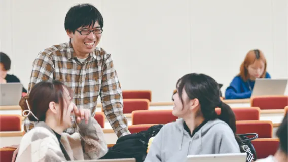 教室で笑顔で女子学生に話しかけているチェック柄のシャツを着た男性