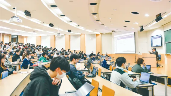 広い講義室でノートPCを開いて前方のスクリーンを見つめる学生たち