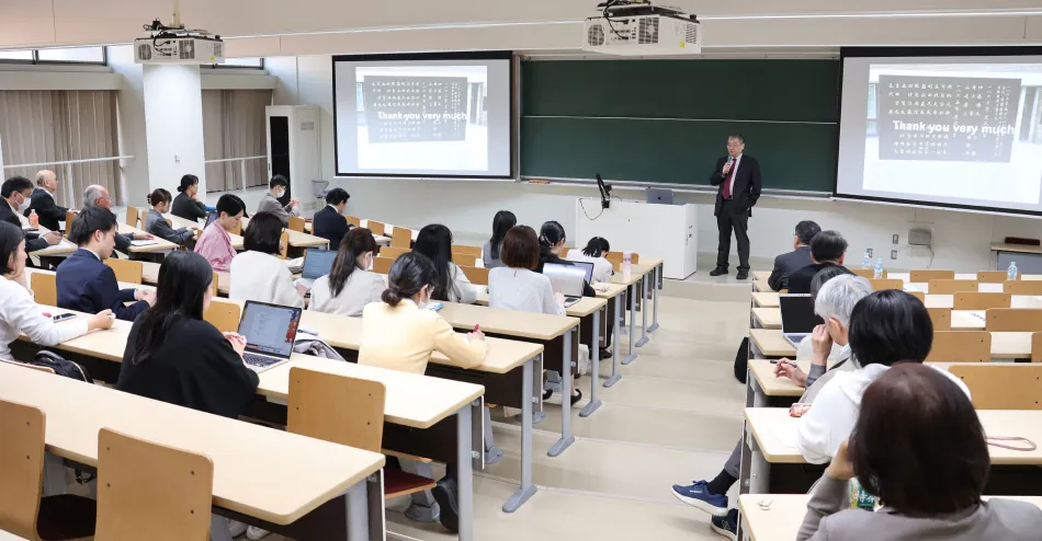 プロジェクター画面の前でマイクを持って講義を行うスーツ姿の講師