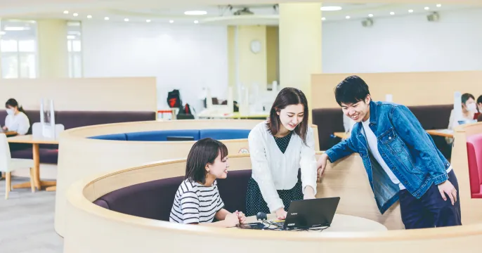 丸いテーブルで、PCを囲み笑顔で話し合う3人の学生たち