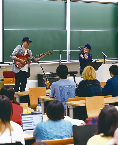 教室でギター演奏を披露する男性と見守る学生たち